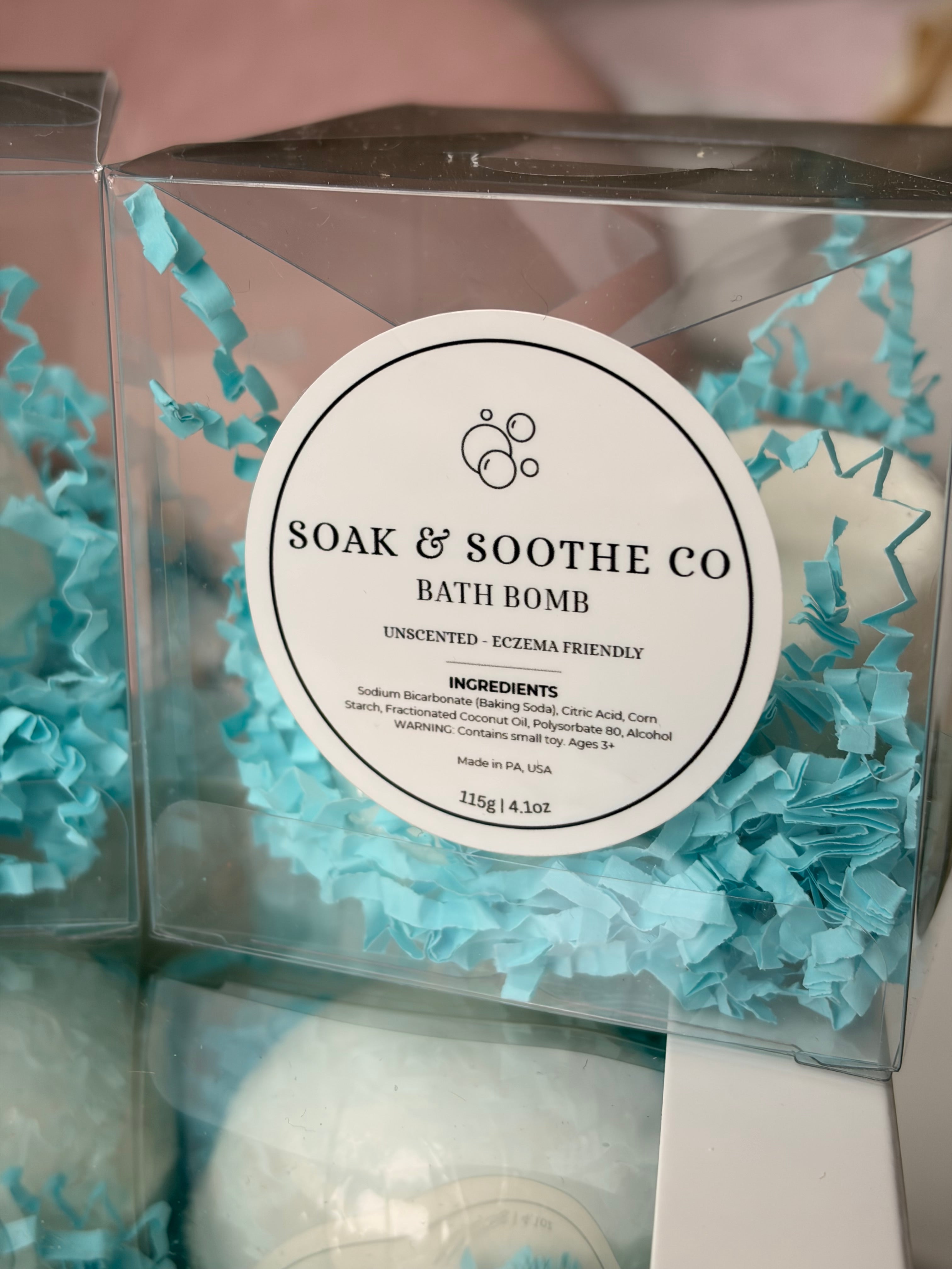 Bath bomb in a clear box with decorative blue tissue paper, branded 'Soak & Soothe Co'.